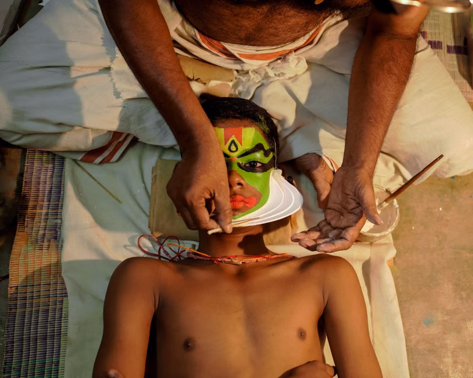 A Kathakali artist lying back while a makeup artist paints his face green