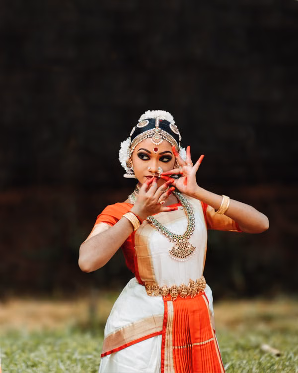 A classical dancer in a white and red costume striking a mudra in an open field at dusk