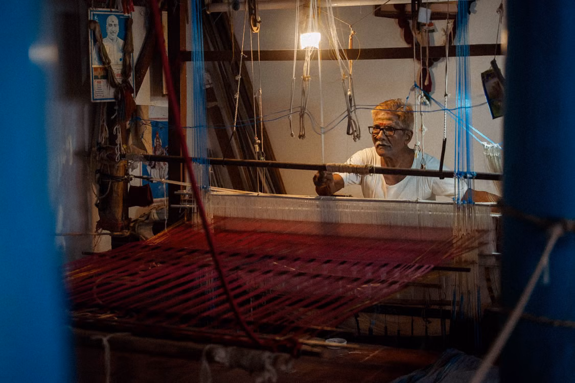 A weaver at a handloom in a workshop, red threads stretched across the frame