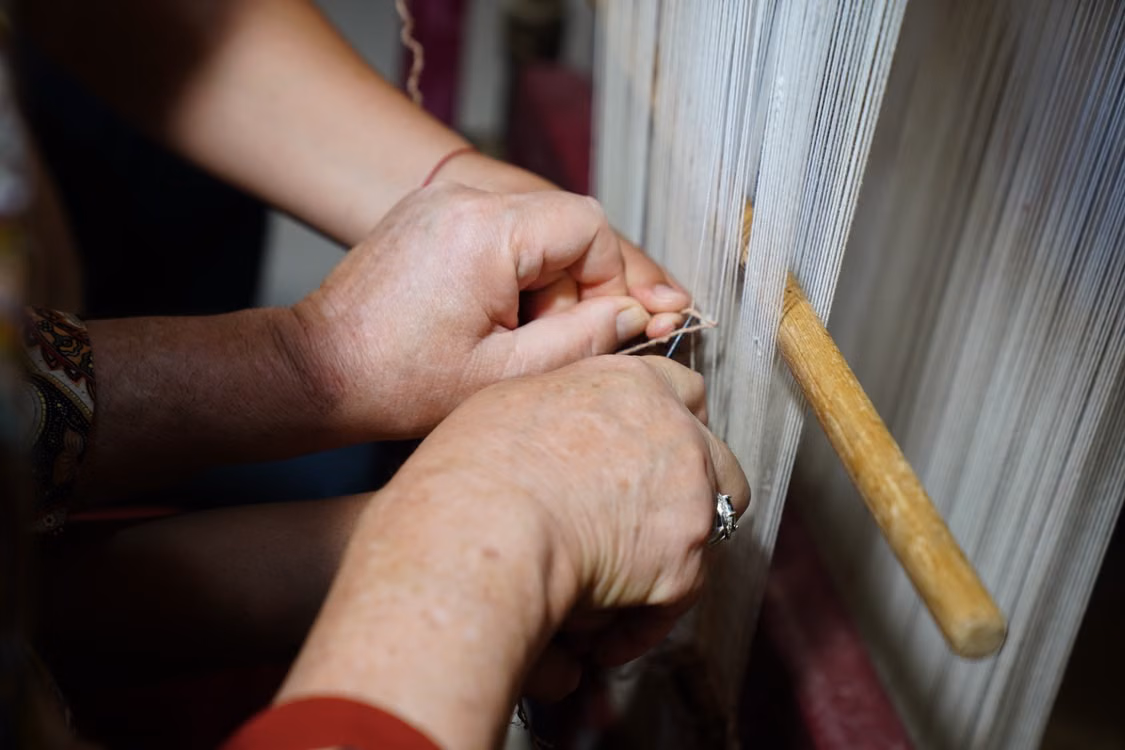 Hands working fine white threads on a loom, a wooden shuttle held in place