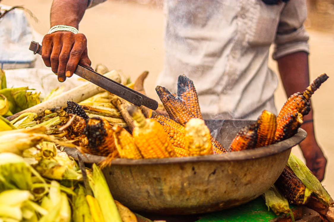 A vendor slicing roasted corn into a wide bowl of fresh produce
