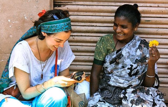 Two women laughing together over a phone, one in a green headscarf, the other holding a yellow flower