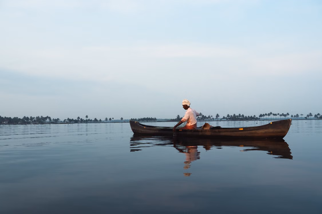 A lone fisherman in a wooden canoe on a calm, hazy backwater
