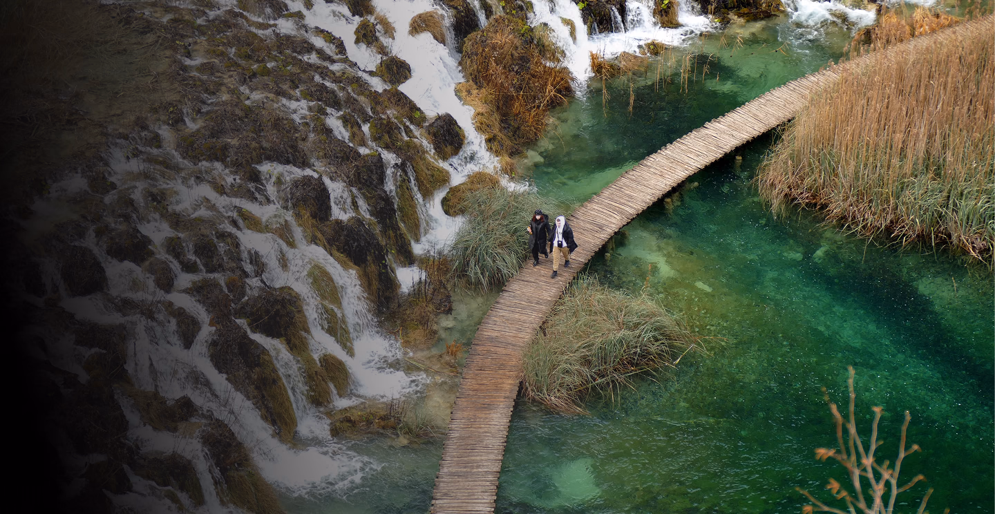 Two travellers crossing a wooden boardwalk above turquoise water, beside a cascade of waterfalls