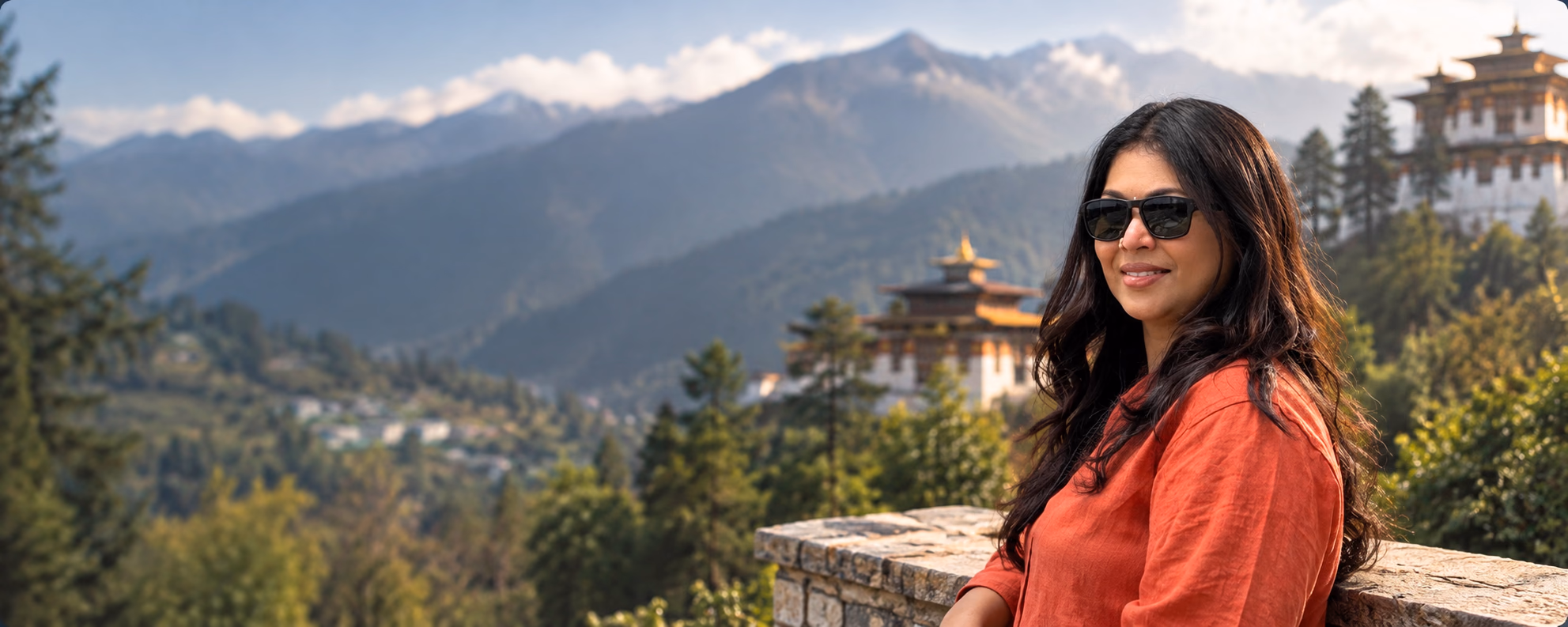 Sreeja Janaki in a rust-red linen shirt and sunglasses, leaning on a stone wall with forested Himalayan mountains and a dzong behind her