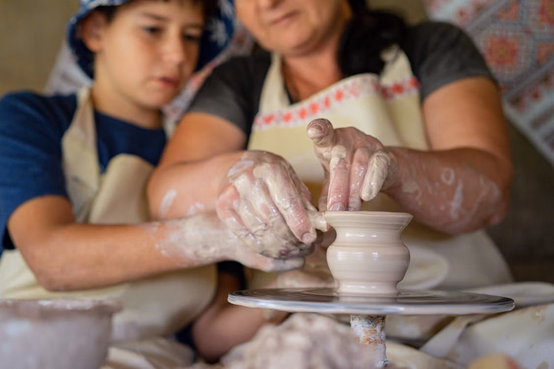 A craftswoman guiding a child’s hands at a pottery wheel
