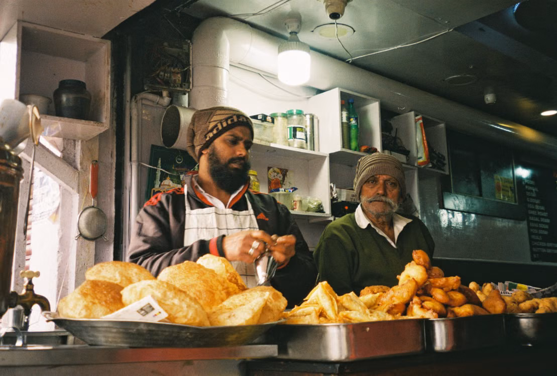 Two cooks at a street food counter piled with fried bread