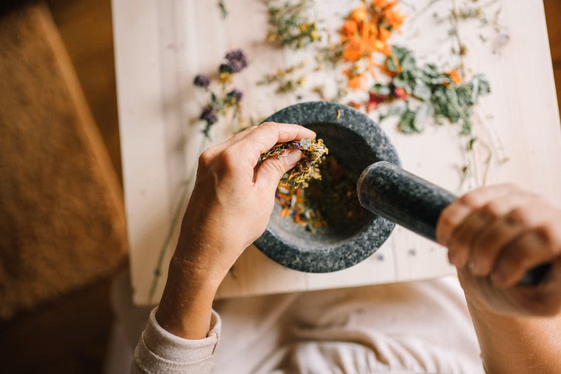Hands grinding herbs in a stone mortar beside dried flowers