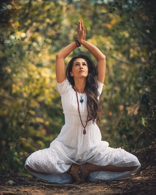 A woman in white with her arms raised in a yoga pose among trees