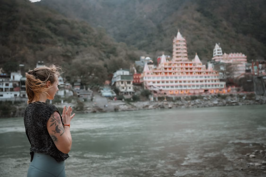 A woman with her hands in prayer beside a river, a temple rising behind her