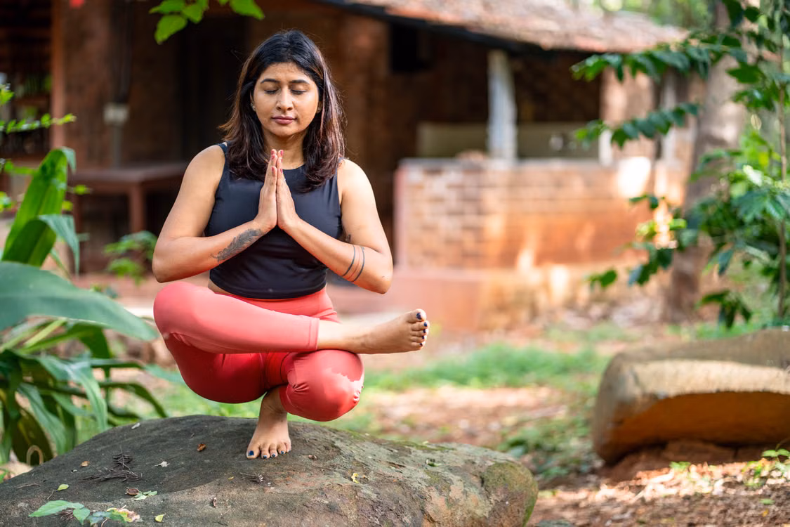 A woman in a prayer squat on a rock in a garden