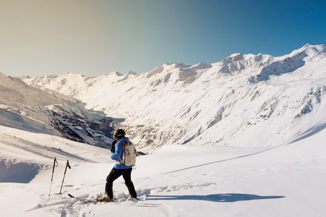 A hiker with poles on a snowy slope below peaks