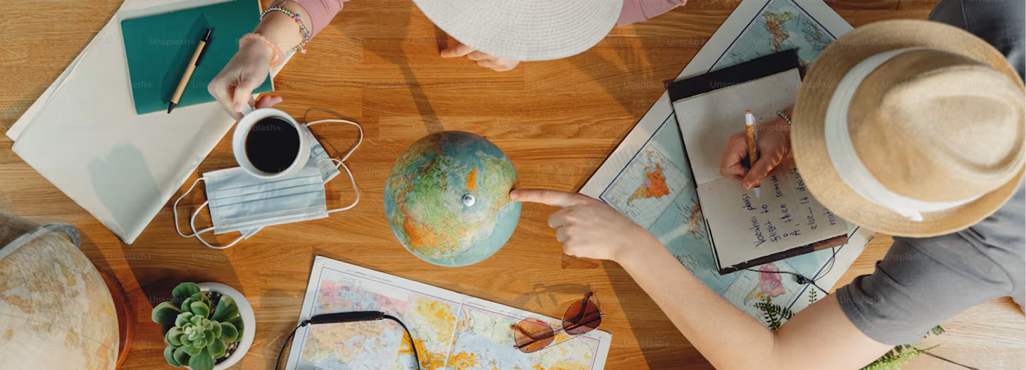 Travellers planning a trip over a wooden table, a hand pointing at a globe beside maps, a notebook and a coffee