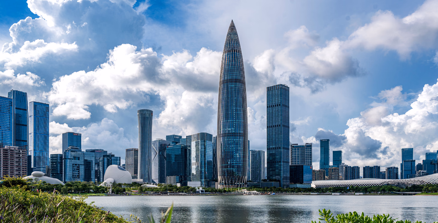 A slender glass skyscraper rising above a modern waterfront skyline under a cloudy blue sky