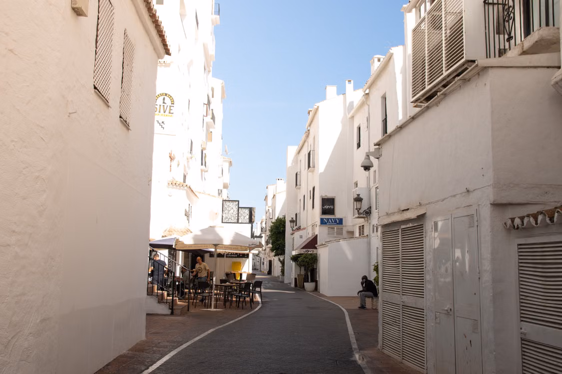 A quiet whitewashed lane with a café terrace and a few people in the shade