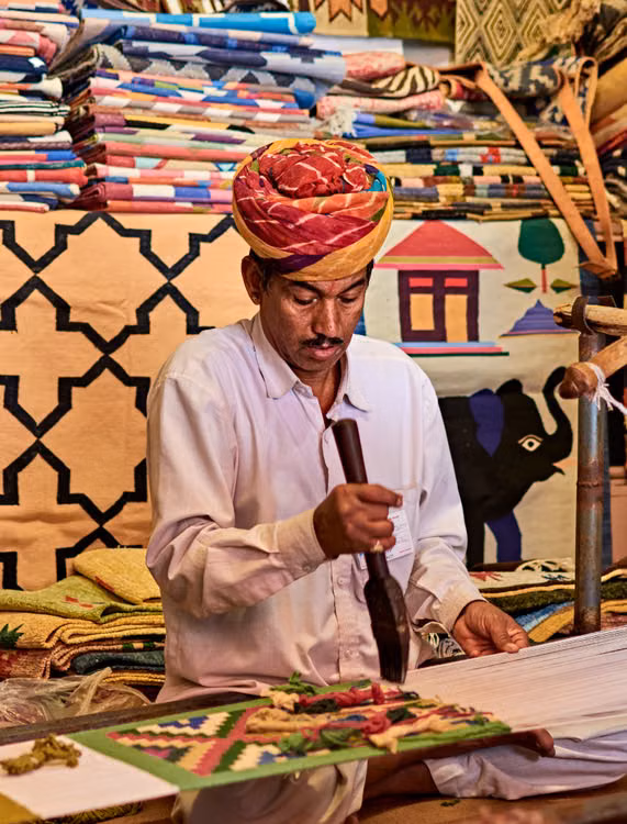 A man in a red turban cutting cloth among stacked textiles