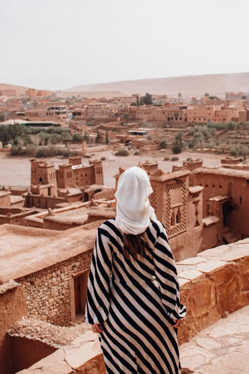 A woman in a striped robe and white headscarf looking out over a clay-walled kasbah
