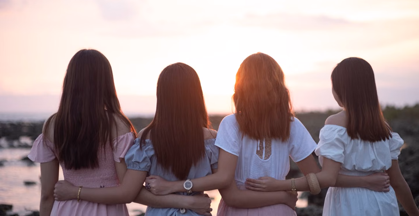 Four women with their arms around each other, seen from behind, looking out over the sea at sunset